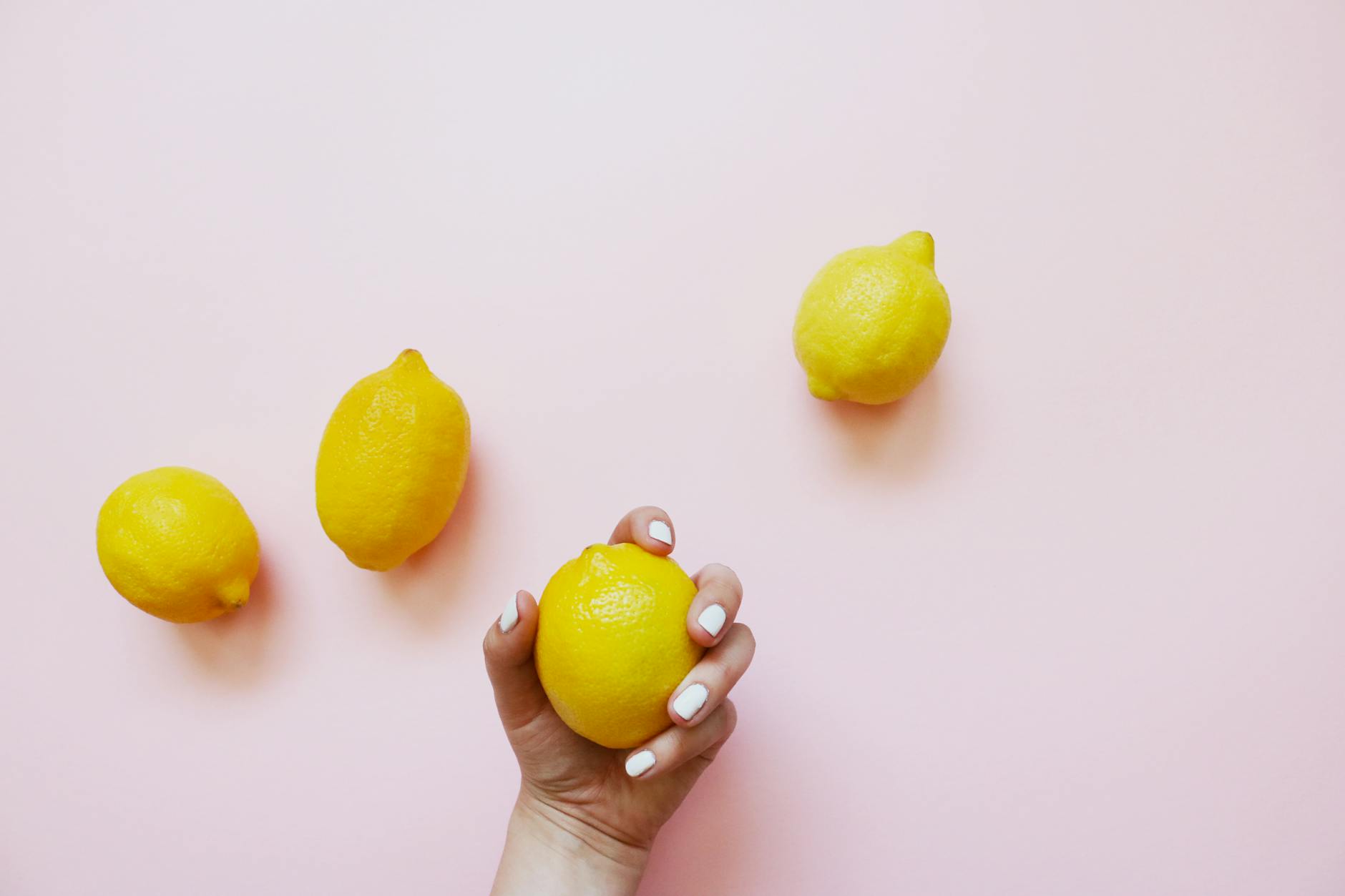 A hand holding a fresh lemon against a soft pink background with additional lemons nearby