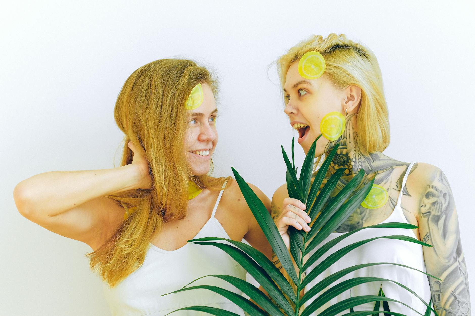 Two smiling women with lemon slices and tropical plant, expressing joy and fun indoors